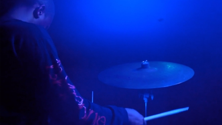 A man with dark skin is playing on a drum kit. The room is dark, with a blue light shining on him.